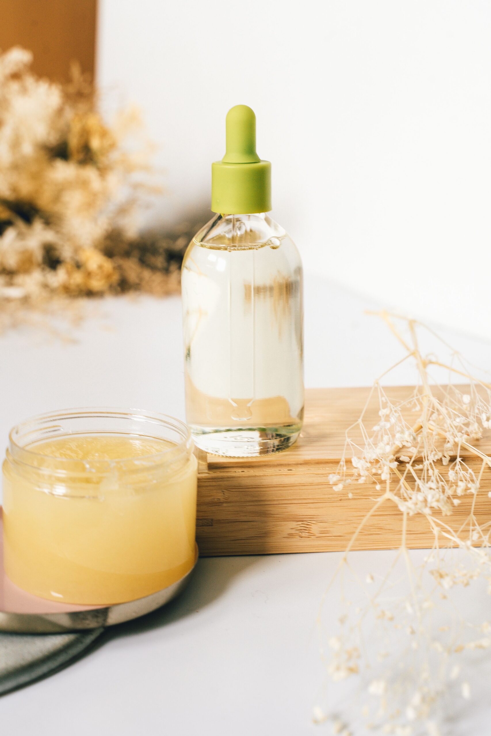 Aesthetic display of a glass bottle and jar of skin care products on a wooden platform with dry flowers.
